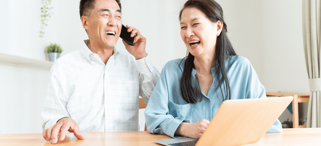ミドル世代の夫婦が笑顔で電話をかけながらパソコンを使っている